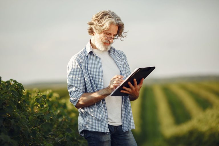 Agricultor idoso anota informações em um caderno enquanto observa uma plantação verde.